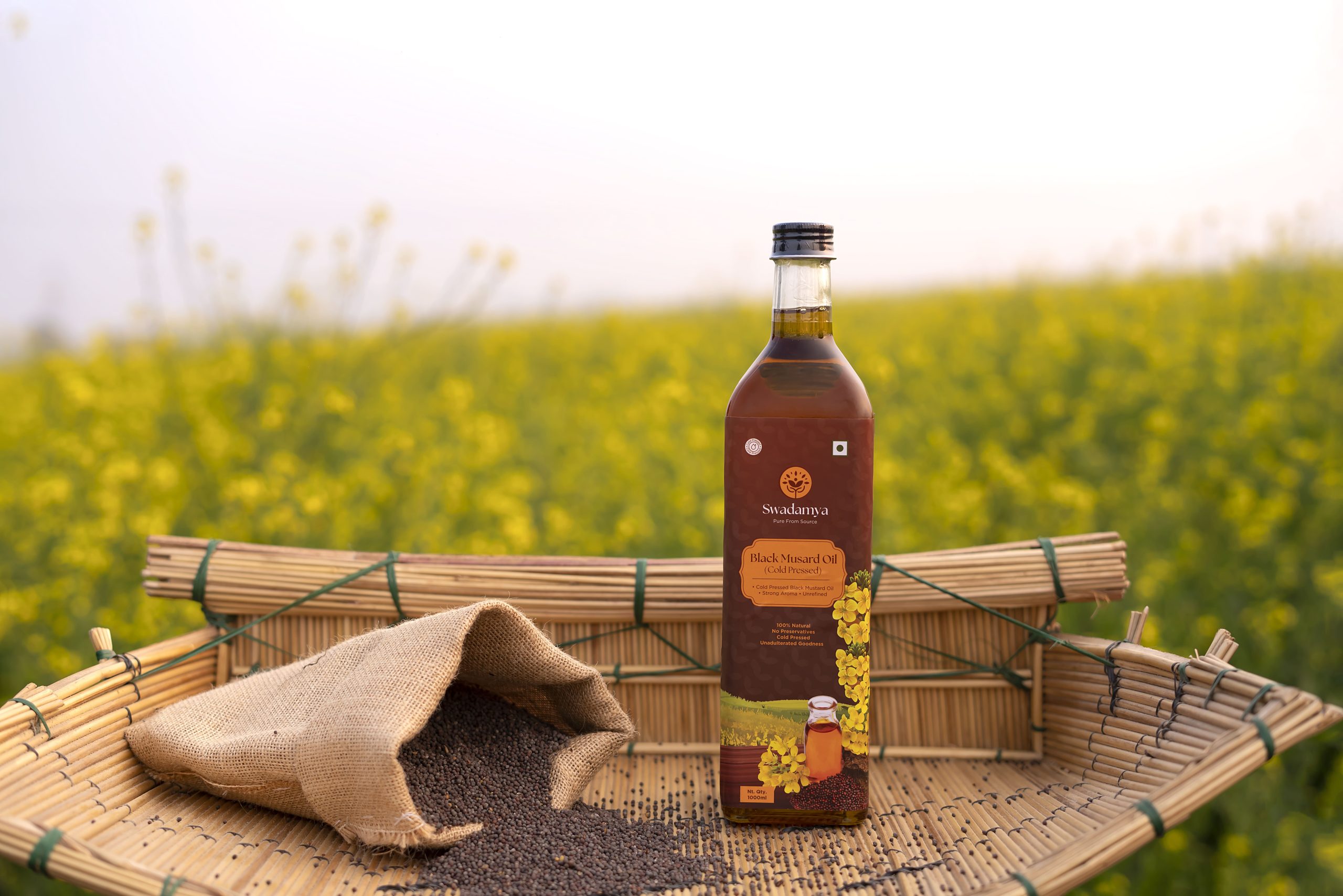 Swadamya cold pressed black mustard oil bottle placed in a natural mustard farm setting